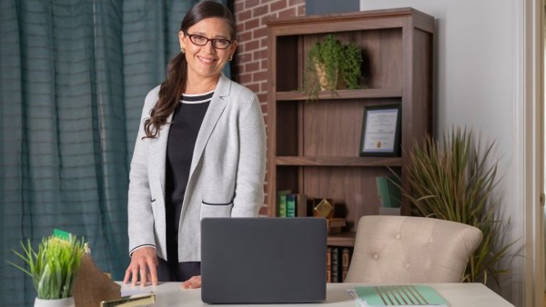 A business woman poses in her nicely furnished office wearing glasses and a blazer.